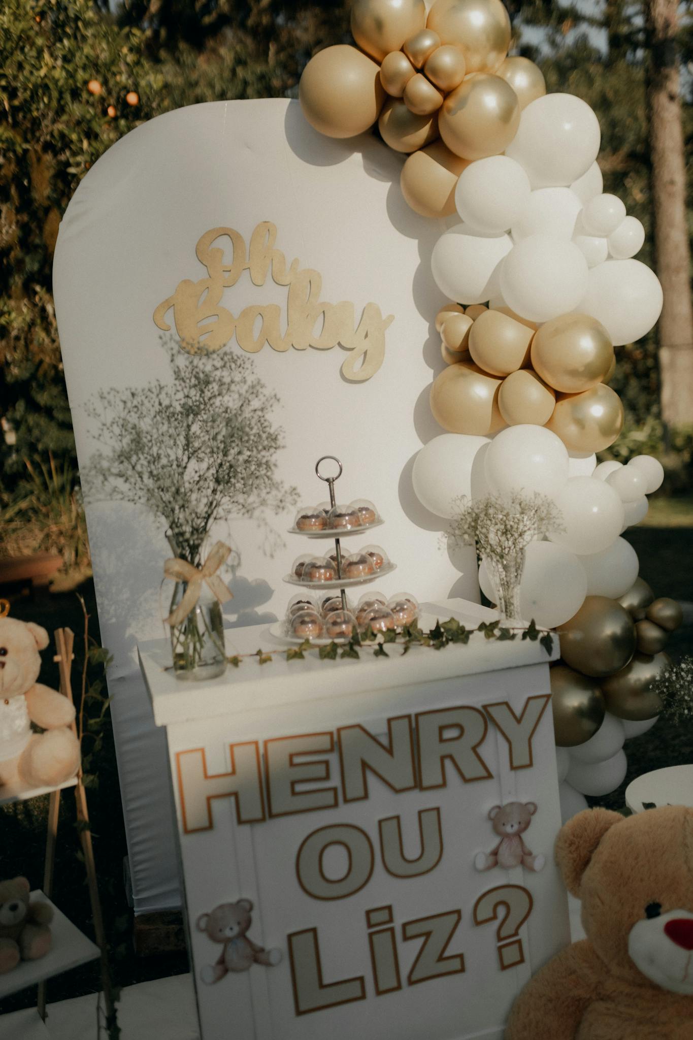 Elegant gender reveal setup with gold and white balloons and teddy bears outdoors.
